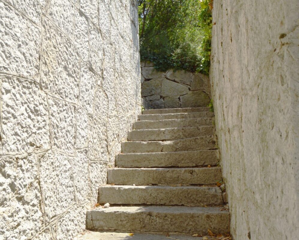 Old narrow staircase with stone wall on both sides, which leads up, illuminated by sunlight